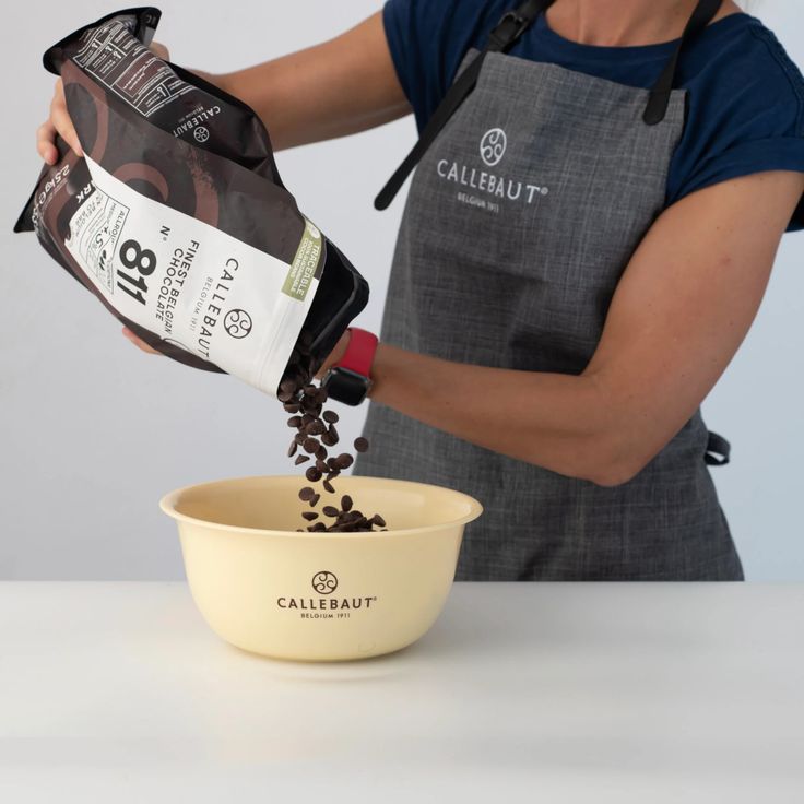 Person pouring chocolate chips from a Callebaut bag into a bowl, wearing a gray apron with the brand logo.